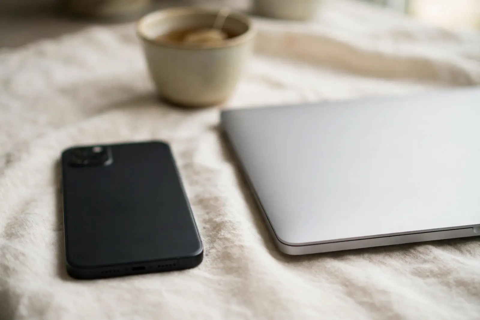 A smartphone lying face down on pale cream linen beside a closed laptop, a ceramic cup of tea out of focus behind